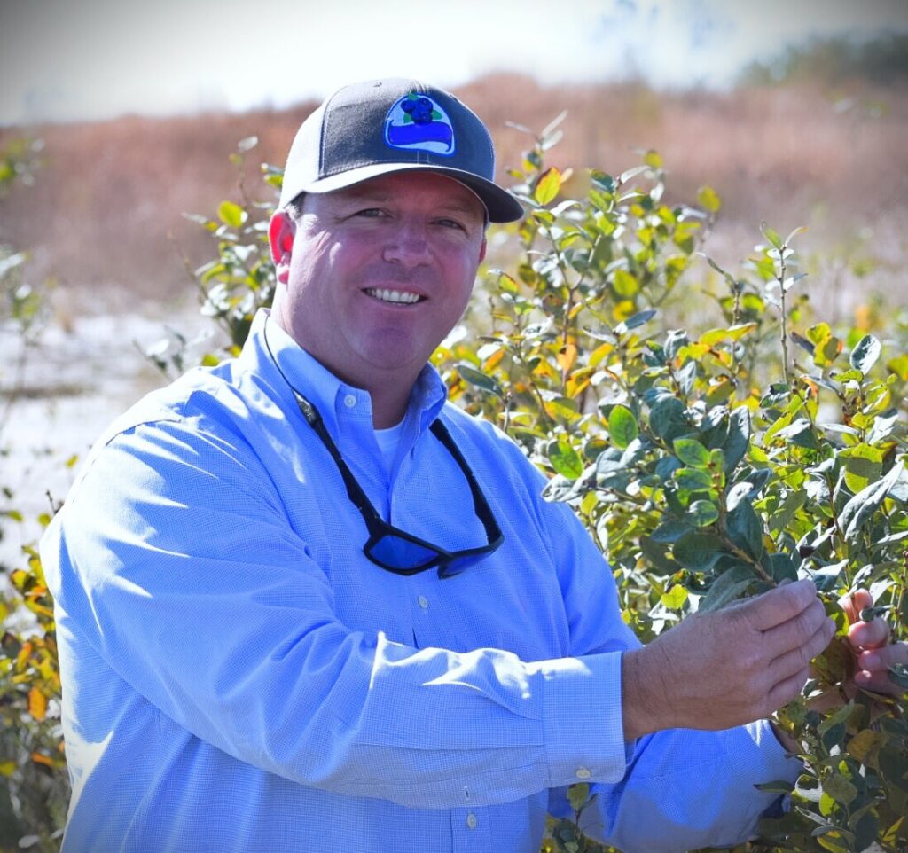 Man in a blue shirt and gray cap smiles while inspecting green leaves on a bush in a sunny outdoor setting.