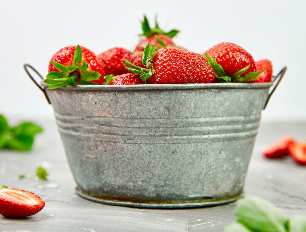 Ripe red strawberries in a rustic metal bowl on a gray table, with fresh strawberries scattered around.